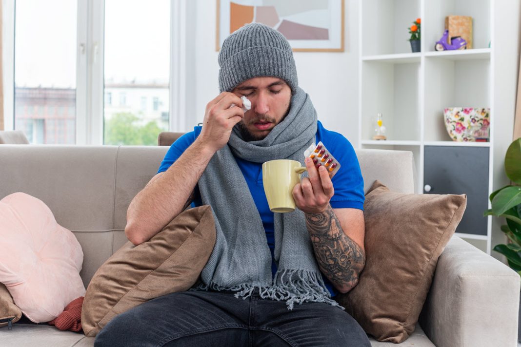 weak-young-ill-man-wearing-scarf-winter-hat-sitting-sofa-living-room-holding-packs-pills-cup-tea-wiping-eye-with-closed-eyes